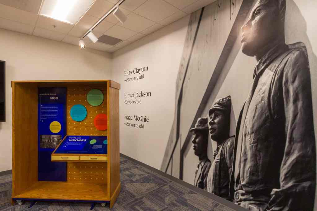 A wood cabinet with signs explaining the Duluth lynchings. On the wall to the right, a large photograph of a memorial for Elias Clayton, Elmer Jackson and Isaac McGhie, the three young African American me who were lynched.
